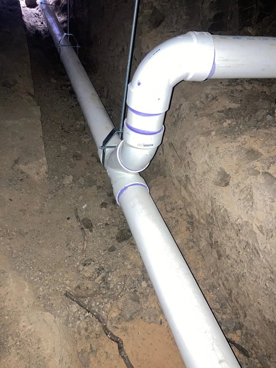 New white PVC drain line hung on straps in a dug-out crawlspace under a house