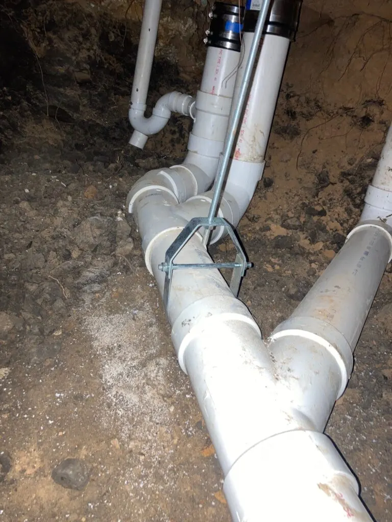 A new PVC wye and drain run under a house, supported by hangers before the tunnel is backfilled.
