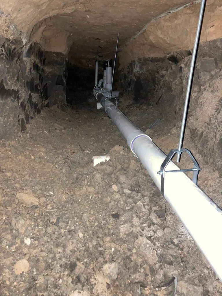 Hand-dug tunnel under a slab home with a new PVC drain line hung along its length.